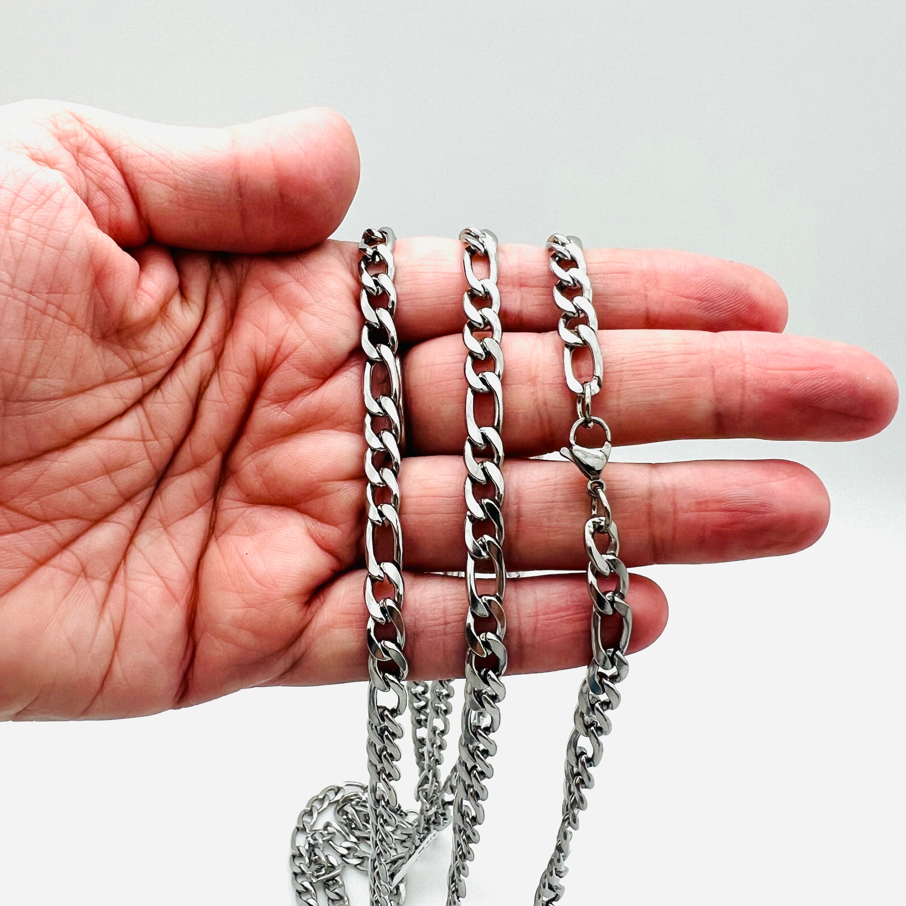 silver tone Figaro chain necklace with lobster claw clasp displayed on a hand with a white background. 