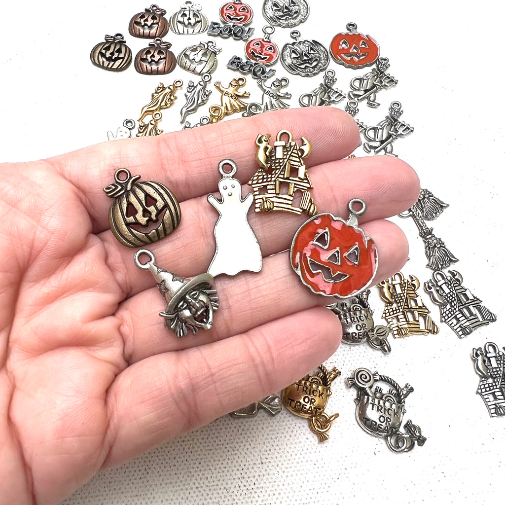 bronze jack-o-lantern charm, white enamel ghost charm, gold haunted house charm, silver witch face charm, orange enamel jack-o-lantern charm in a hand for scale on a white background