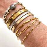 an arm featuring 9 stacked bracelets and bangles, including a golden paperclip chain bracelet with a white background. 
