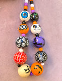 painted wooden beads with a Halloween theme. Beads include an eyeball, the word "boo", a scream face, spiderweb, jack-o-lantern faces, a witch on a broom and a black cat face. Beads are paired with purple, orange, black and white beads