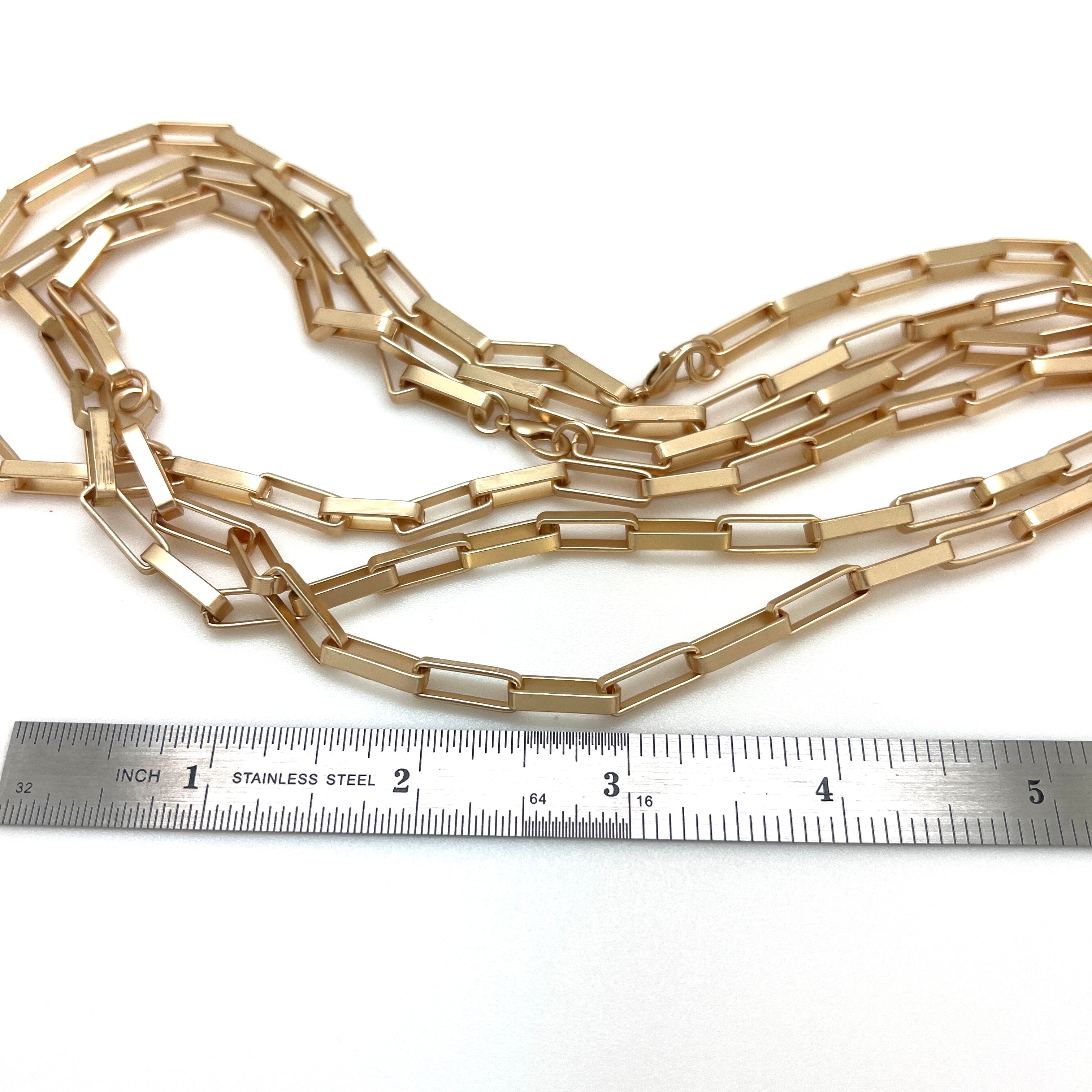 multiple matte finished, golden large square paperclip link necklace with lobster claw clasp displayed above stainless steel ruler on white background. 