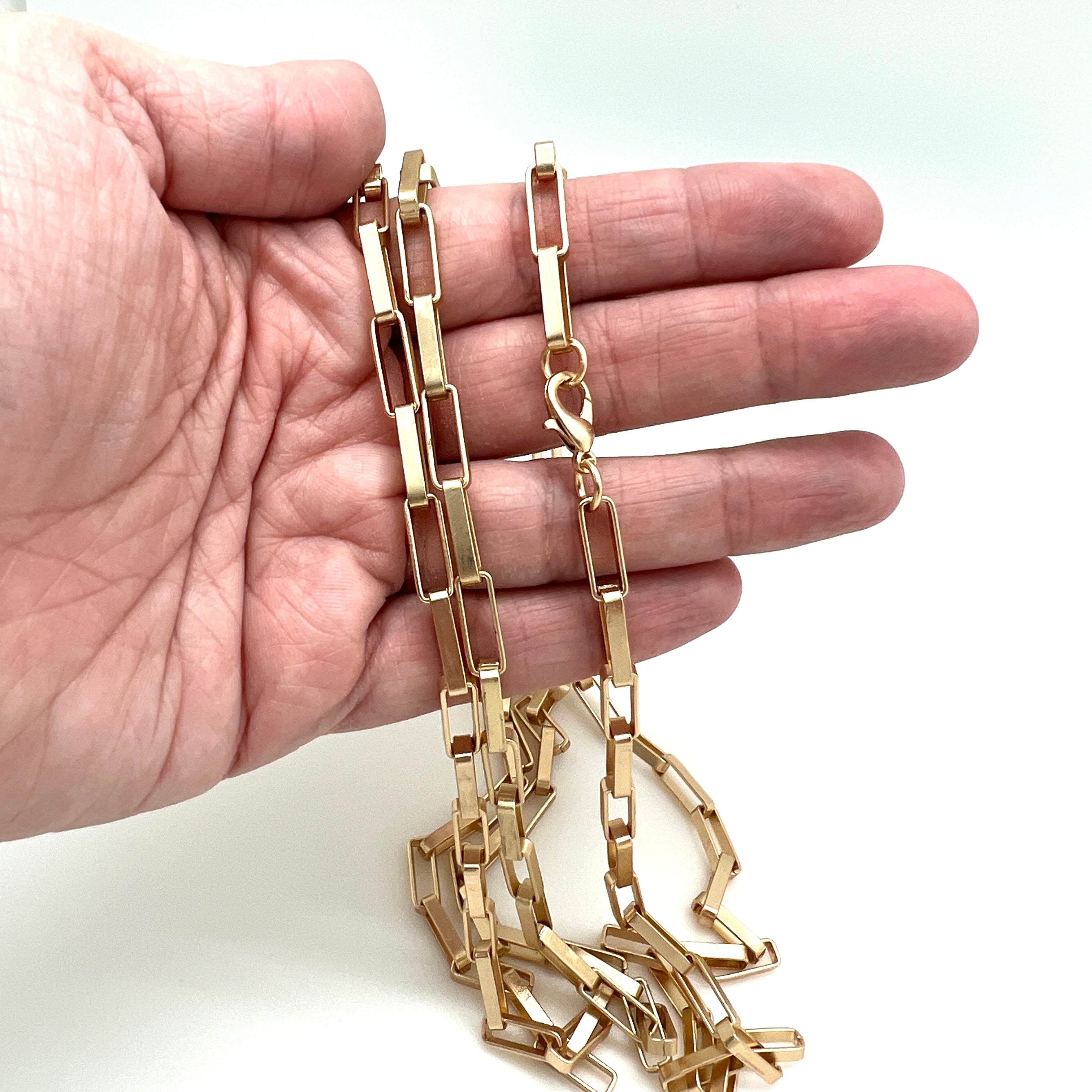 multiple matte finished, golden large square paperclip link necklace with lobster claw clasp draped over a hand against white background. 